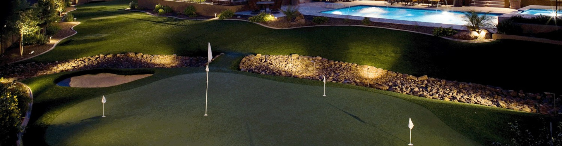 Aerial night view of a synthetic turf putting green with four flag pins, boulder and sand bunkers, landscape lighting, and a resort-style pool in a luxury backyard